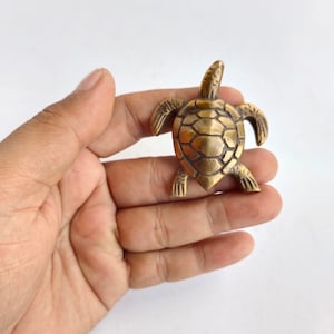 May include: A small, gold-colored metal turtle figurine. The turtle is facing forward with its head raised and its legs outstretched. The turtle's shell is detailed with a textured pattern.