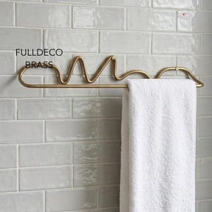 May include: A brass towel rack with a wavy, continuous line design mounted horizontally. The metal frame features a smooth, matte finish and includes the text FULLDECO BRASS. A plain white rectangular towel hangs over the lower horizontal bar.