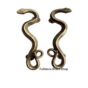 Op de afbeelding: Een paar messing deurgrepen in de vorm van slangen. De grepen zijn in een gekrulde positie, met de slangenkoppen naar boven gericht. De grepen zijn goudkleurig en hebben een getextureerd oppervlak.