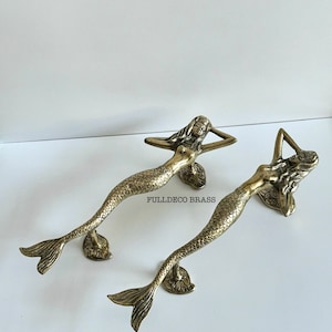 May include: Two gold mermaid door handles with a detailed design. The mermaids are lying on their sides with their tails curled up behind them. The handles are made of solid brass and have a polished finish.