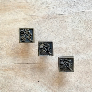 May include: Three antique brass cabinet knobs with a leaf design. The knobs are square shaped and have a slightly raised design.