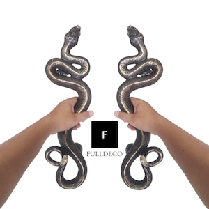 Könnte beinhalten: Zwei schwarze und goldene Schlangentürgriffe. Die Griffe haben die Form einer Schlange mit dem Kopf nach vorne und dem Schwanz um den Griff gewickelt. Die Griffe werden von zwei Händen gehalten.