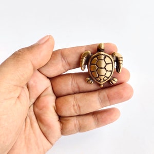 K&ouml;nnte beinhalten: Eine kleine, goldfarbene Schildkr&ouml;tenfigur mit einer detaillierten Schale. Die Schildkr&ouml;te sitzt auf der Hand einer Person.