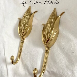 May include: Two gold corn cob hooks. The hooks are shaped like corn cobs with a textured surface. The text "2x Corn Hooks" is written above the hooks.