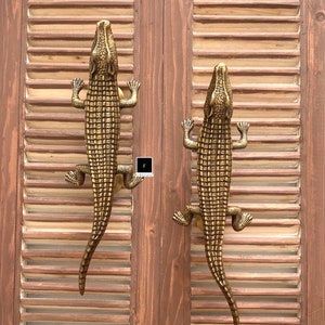 May include: Two gold-toned alligator door handles with detailed scales and open mouths. The handles are mounted on a wooden door with brown painted louvered panels.
