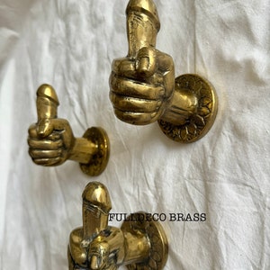 May include: Three gold-toned metal hooks shaped like hands giving a thumbs up, with the thumb being a phallic symbol. The hooks are mounted on a white wall.