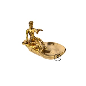May include: A gold-colored metal sculpture of a man sitting on a rock, holding a small object in his hand. The sculpture is on a gold-colored metal tray.