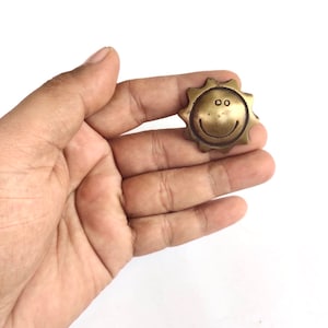 May include: A small, gold-coloured metal sun with a smiling face. The sun has a textured surface and is held in a hand.
