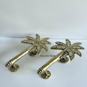 Puede incluir: Dos tiradores de puerta de palmera dorada con acabado cepillado. Los tiradores están hechos de latón macizo y presentan un diseño detallado de palmera. Los tiradores están montados en una base redonda con dos tornillos.