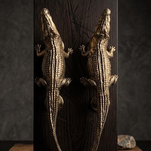 Könnte beinhalten: Zwei goldfarbene, metallene Türgriffe in Alligatorform, die vertikal auf einer dunkelbraunen Holzplatte montiert sind. Jeder Griff hat eine strukturierte, schuppige Oberfläche und ist mit einer runden Basisplatte in der Mitte befestigt.