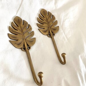 May include: Two gold-toned metal hooks shaped like tropical leaves. The hooks are mounted on a white surface.