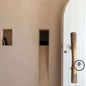 May include: A brass door handle with a geometric pattern. The handle is attached to a white door with a white trim. The door is in a room with white walls and two rectangular niches.