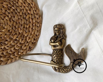 May include: Gold-toned mermaid-shaped toilet paper holder with a seashell in her hand. The holder is mounted on a white wall.