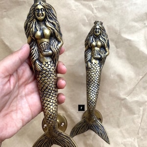 Może przedstawiać: Dwa złote uchwyty do drzwi w kształcie syreny z koroną i muszlą w dłoniach. Ogon syreny jest ozdobiony łuskami.