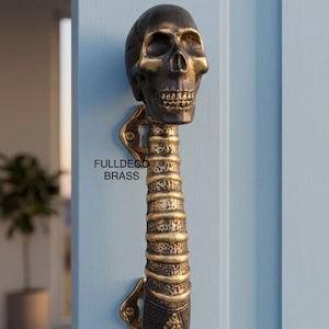 May include: A brass door handle featuring a detailed human skull at the top and a spinal column design extending downwards. The handle is mounted on a light blue door with visible screws at the top and bottom brackets. The text FULLDECO BRASS is visible.