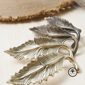 Könnte beinhalten: Drei goldene, blattförmige Haken mit einem detaillierten Design. Die Haken sind in einer Reihe angeordnet, wobei der größte Haken vorne und der kleinste Haken hinten ist. Die Haken befinden sich auf einer hellbraunen Holzoberfläche.