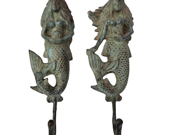May include: Two bronze-effect mermaid wall hooks. Each hook depicts a mermaid with flowing hair and a fishtail. The mermaids are in a standing pose. The hooks are curved and extend downwards. The hooks are made of metal.