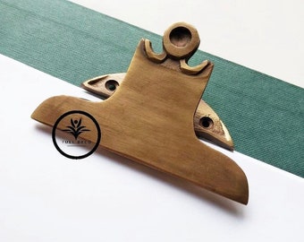 Puede incluir: Una pinza de portapapeles de latón con un diseño decorativo. La pinza tiene la forma de una letra 'S' estilizada con una parte superior redondeada. La pinza está sobre una superficie blanca.