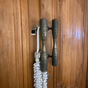 Może przedstawiać: Para ozdobnych, mosiężnych klamki drzwiowych z misternymi geometrycznymi wzorami, zamontowanych na drewnianych drzwiach z rzeźbionymi detalami. Biały frędzl z koralików zwisa z klamki.