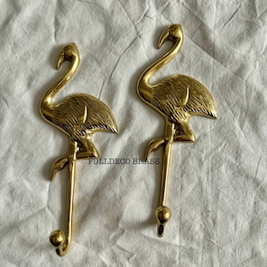 Może przedstawiać: Dwa złote haczyki w kształcie flaminga z fakturowanym wykończeniem. Haczyki są przymocowane do białego tekstylnego tła. Na tle widoczny jest tekst "FULLDECO BRASS".