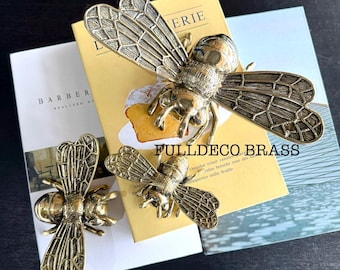 Peut inclure: Trois figurines d'abeilles en métal doré avec des ailes et des corps détaillés. La plus grande abeille est sur un livre avec le texte "FULLDECO BRASS" et "ERIE".