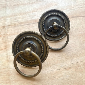 Puede incluir: Dos tiradores de puerta de latón antiguo con un diseño de cuerda alrededor del borde. Cada tirador tiene una manija redonda en forma de anillo.