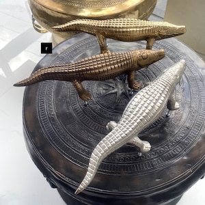 May include: Three decorative crocodile figurines, one silver, one gold, and one bronze, are displayed on a round, dark gray surface with intricate patterns. The crocodiles are positioned in a circle, with their tails pointing outwards.