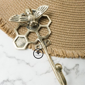 May include: Gold metal bee and honeycomb hook. The hook is attached to a honeycomb design with a bee on top. The hook is on a white marble surface.