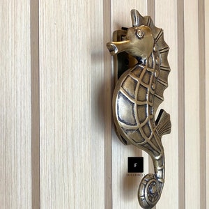 May include: A gold-toned seahorse door knocker with intricate details. The knocker is mounted on a wooden door with vertical panels.