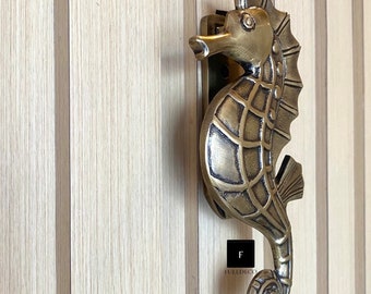 May include: A gold-toned seahorse door knocker with intricate details. The knocker is mounted on a wooden door with vertical panels.