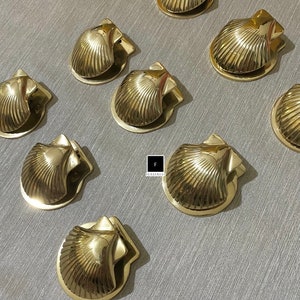 May include: A set of eight gold-toned metal seashell-shaped paperweights. Each paperweight has a detailed seashell design and a spring-loaded clip.