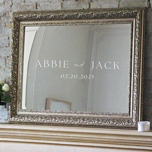 May include: A silver ornate framed mirror with a white inscription that reads "Abbie and Jack 02.20.2025".