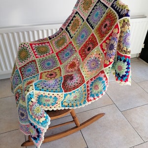 May include: A colourful crocheted blanket with a floral pattern, draped over a wooden rocking chair. The blanket is made of various shades of pink, blue, green, yellow, and red.