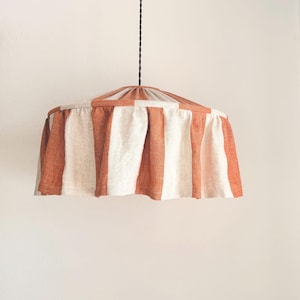 May include: A pendant light fixture with a striped lampshade. The shade features alternating vertical stripes of burnt orange and off-white. The light hangs from a black cord against a plain white background. The shade has a gathered top.