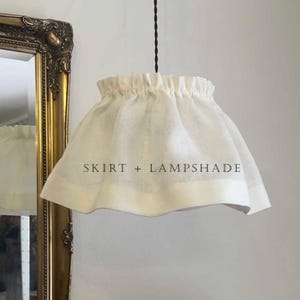 Op de afbeelding: Een witte stoffen lampenkap met een geplooide bovenrand en een brede zoom, opgehangen aan een zwart gedraaid snoer. De tekst SKIRT + LAMPSHADE is in het zwart in het midden van de kap gedrukt. Het ontwerp heeft een uitlopend, rokachtig silhouet.