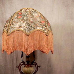 May include: A vintage-style lampshade with a floral pattern in shades of green, brown, and pink. The lampshade has a scalloped edge with a fringe trim in a peachy-orange color.
