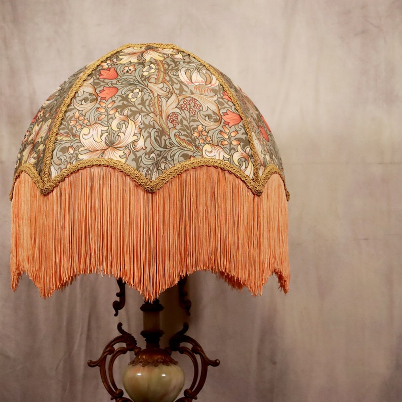 May include: A vintage-style lampshade with a floral pattern in shades of green, brown, and pink. The lampshade has a scalloped edge with a fringe trim in a peachy-orange color.