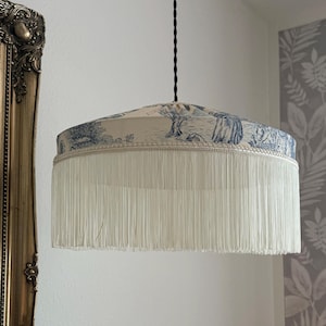 Puede incluir: Pantalla de lámpara colgante con flecos color crema y un estampado toile de Jouy azul y blanco. La pantalla tiene forma cilíndrica con un ribete decorativo en la parte superior. El cable es negro.