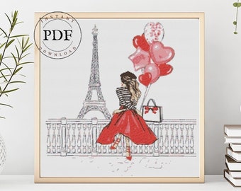 May include: A cross-stitch pattern featuring a woman in a red dress and a striped shirt holding balloons in front of the Eiffel Tower. The pattern is framed and displayed on a white shelf with a potted plant and a stack of books.