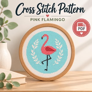 May include: A circular wooden embroidery hoop containing a cross stitch pattern of a pink flamingo on a light blue background, framed by white leaf sprigs. The text Cross Stitch Pattern PINK FLAMINGO and a red circular PDF icon are visible.