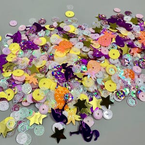 May include: A collection of loose sequins in various shapes and colors, including yellow circles, gold stars, orange pineapples, and purple flamingo silhouettes, mixed with iridescent, white, and pink circular sequins of different sizes.