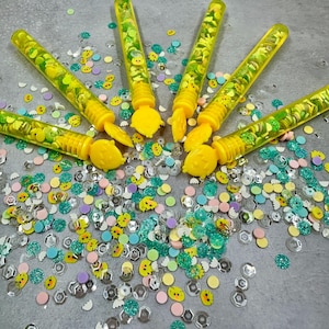 May include: Six yellow plastic bubble wands filled with colorful confetti and glitter. The wands are arranged in a circle on a gray surface.