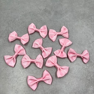 May include: A collection of light pink satin bows arranged on a gray surface. Each bow is tied with a classic bow shape, showcasing a delicate and feminine aesthetic. The bows are evenly spaced, creating a visually appealing pattern.