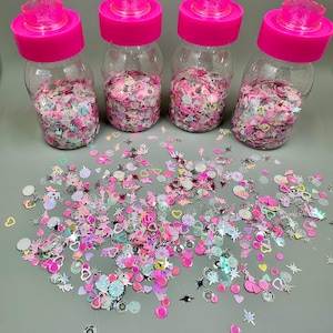 May include: Four clear plastic jars filled with pink and iridescent confetti. Each jar has a bright pink lid. Confetti includes hearts, stars, and flower shapes. Some confetti is scattered on a gray surface.