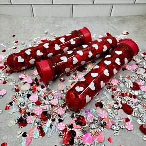 May include: Three red plastic tubes filled with red and white heart confetti. The tubes have a white cap on one end and a red cap on the other end. The tubes are sitting on a surface covered in red, white, and pink confetti.