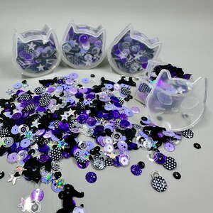 May include: A collection of loose sequins in shades of purple, black, and white, featuring star, cat, and circular shapes with polka dot and holographic finishes. Four clear, cat-shaped plastic containers are included, some filled with the sequins.