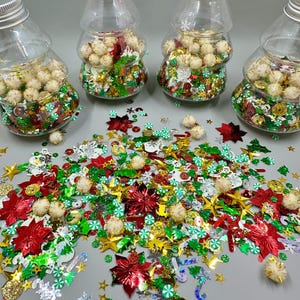 Op de afbeelding: Vier heldere, kerstboomvormige flessen met zilveren deksels, gevuld met feestelijke confetti en kleine, witte pompons. Verspreid liggen rode poinsettia's, gouden sterren, groene bomen en sneeuwvlokvormige pailletten. Een knutselbenodigdheid met vakantiethema.