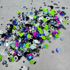 May include: A variety of colorful and sparkly Halloween-themed embellishments, including ghosts, stars, eyes, and moons. The embellishments are scattered on a gray surface.