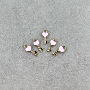 May include: Five gold-colored key charms with pink heart-shaped tops. The charms are arranged in a semi-circle on a gray surface.