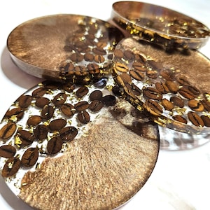 May include: Four round resin coasters with coffee beans and gold flakes embedded in them. The coasters have a clear resin base with a bronze-colored swirl design. The coffee beans are a dark brown color.